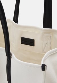 Sac fourre-tout en toile blanche avec des poignées noires et une étiquette intérieure cousue avec le nom de la marque "Emporio Armani."