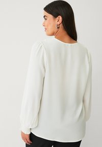 Hvid bluse med lange, puffede ærmer og rund halsudskæring. Glat, letvægtsstof; samlet detalje ved ærmeafslutninger.