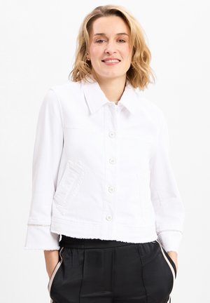 Frau lächelt, trägt eine weiße geknöpfte Jacke mit Dreiviertelärmeln und schwarze Hose mit weißen Seitenstreifen, die Hände in den Taschen.