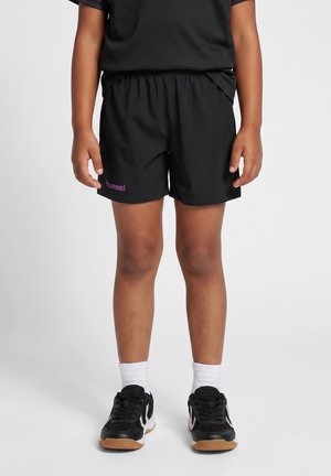 Barn iført sorte sportsshorts med lilla logo, sorte sneakers, hvide sokker og en sort t-shirt, stående mod en grå baggrund.