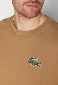 Hombre vistiendo una camiseta marrón de cuello redondo con un logo de cocodrilo bordado en verde en el lado izquierdo del pecho y con barba corta visible.