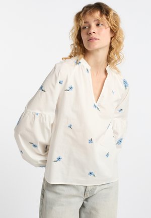 Femme portant un chemisier blanc ample avec de petites fleurs bleues et des manches ballon, associé à un jean en denim bleu clair.