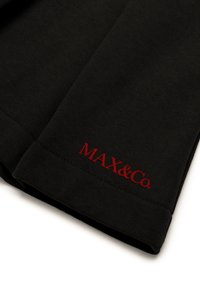 Sort stof med en glat tekstur, der har et rødt "MAX&Co." logo broderet ved kanten. Designet inkluderer en ren og enkel form.