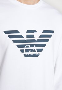 Bílá dlouhá bavlněná košile s dlouhými rukávy, zdobená tmavě šedým pruhovaným logem orla a středně umístěným nápisem "GA" pod logem. Hladká textura, kulatý výstřih.