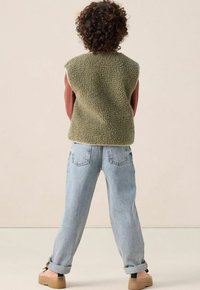 Grüne, flauschige Fleece-Weste über einem braunen Langarmshirt; kombiniert mit hellblauen, locker sitzenden Jeans und beigen Plateauschuhen.