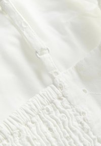 Tissu blanc avec bretelle réglable et délicate broderie florale sur une section froncée d'un vêtement léger.