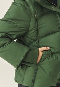 Jachetă puf verde cu un design matlasat, manșete ribbate și un buzunar lateral. Suprafața pare netedă și rezistentă la apă.