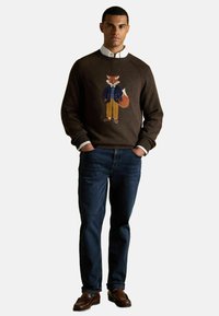 Brauner Wollpullover mit gesticktem Fuchs in blauer Jacke, kombiniert mit blauen Jeans und braunen Mokassins. Weißer Hemdkragen sichtbar.