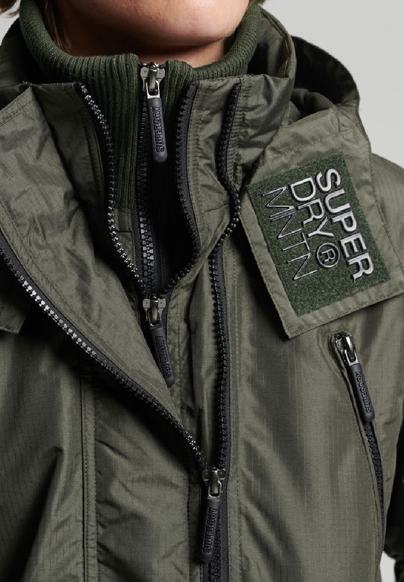 Nahaufnahme einer Person, die geschichtete olivgrüne Jacken mit mehreren schwarzen Reißverschlüssen und einem "SUPER DRY® MNTN"-Aufnäher auf der Brust trägt.