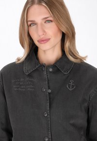 Fekete farmerdzseki, gallérral, gombos záródással és hímzett horgony részlettel. Finom, hasonló színű szöveges dizájnt tartalmaz.