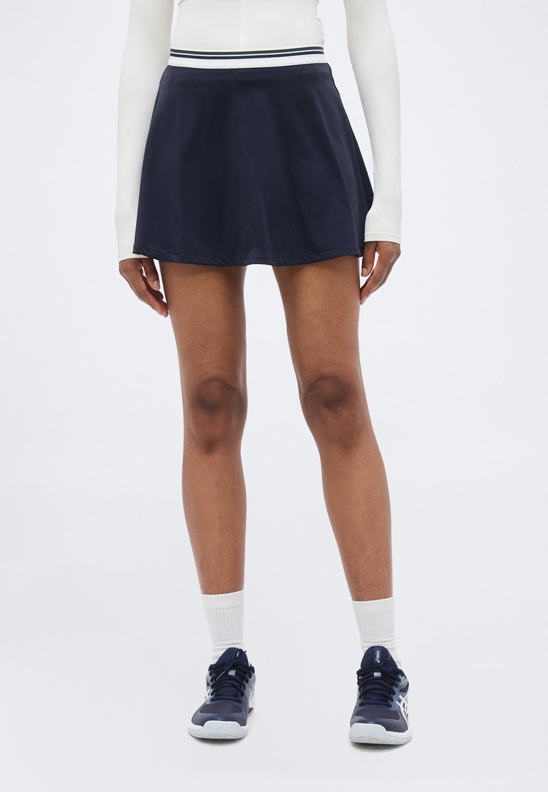 Kvinne iført en marineblå atletisk skjørt med hvitstripet midjebånd, hvit langermet topp, hvite sokker og marineblå joggesko som står.