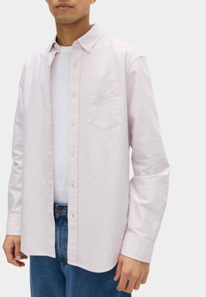 Mand iført en lyserød og hvid stribet skjorte med knapper over en hvid T-shirt med blå jeans, stående mod en ensfarvet baggrund.