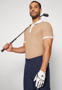 Gaišbrūns īsām piedurknēm polo krekls ar baltu apkakli un piedurknēm, apvienots ar tumši ziliem biksēm. Persona tur golfu nūju un valkā baltu golfu cimdu.