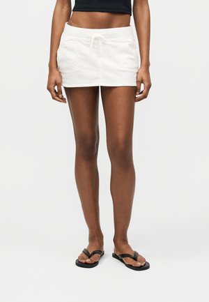 Kvinde iført hvid mini-nederdel med snøre, sort crop top og sorte klipklappere, stående mod en ensfarvet hvid baggrund.