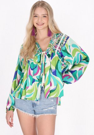 Jonge vrouw draagt een kleurrijke blouse met lange mouwen met een groen, blauw en paars patroon, spijkershorts en paarse kwastoorbellen, glimlachend.