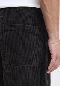 Pantaloni neri in velluto a coste con righe verticali, tasca laterale e vestibilità comoda; la texture del tessuto è morbida e leggermente lucida.