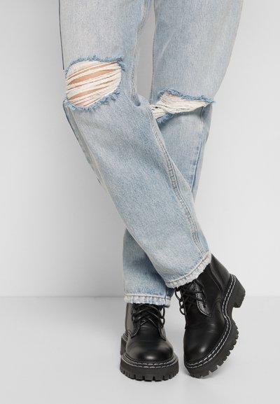 Botas pretas de pele com atacadores, com solas grossas e robustas, emparelhadas com jeans descoloridos azul claro, apresentando rasgos desgastados nos joelhos e fios brancos visíveis.