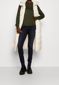 Lång, krämfärgad pufferväst över en mörkgrön topp, kombinerad med smala navy jeans och svarta snörboots, vilket visar en avslappnad stil.