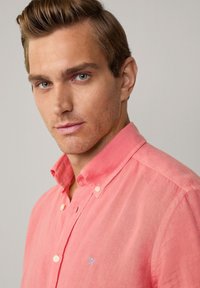 Camicia button-up rosa chiaro con un piccolo logo ricamato blu. Realizzata in un tessuto testurizzato, presenta un colletto e dettagli dei bottoni visibili.