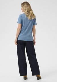 T-shirt en coton bleu clair à manches courtes, associé à un pantalon large bleu foncé. Le mannequin a les cheveux ondulés et se tourne légèrement sur le côté.