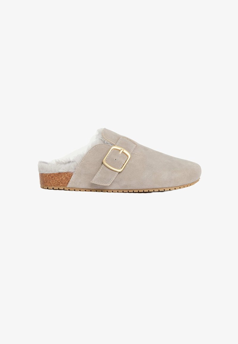 Scarpa slip-on in suede beige con morbida fodera interna, un accento di fibbia dorata e una suola in sughero texturizzata. Esterno liscio con punta arrotondata.