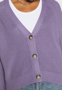 Lila gestrickte Strickjacke mit V-Ausschnitt, drei großen runden Knöpfen und geripptem Saum. Getragen über einem weißen Oberteil mit Rundhalsausschnitt.