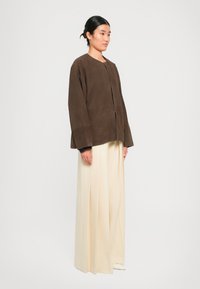 Femme debout portant une veste en daim marron foncé ample et un pantalon large plissé crème devant un fond blanc uni.