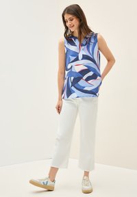 Top blu senza maniche con un motivo geometrico a onde in diverse tonalità di blu, accentuato da linee rosse e bianche. Abbinato a pantaloni bianchi tagliati e scarpe da ginnastica.