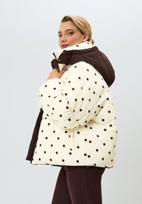Prešívaná bunda z krémovej látky s tmavohnedými bodkami, kontrastnou hnedou kapucňou a zipsovým zapínaním. Elegantný, zaoblený strih.