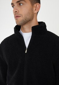 Schwarze Fleecejacke mit hohem Kragen, Reißverschluss und strukturiertem Oberflächenmaterial. Getragen über einem weißen Rundhalsausschnittshirt.