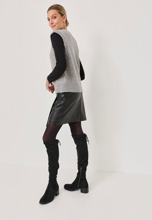 Maglione grigio senza maniche sopra una maglietta nera a maniche lunghe, abbinato a una gonna nera in ecopelle e stivali alti in suede neri.