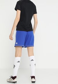 Camiseta deportiva negra, pantalones cortos azules con rayas blancas, calcetines blancos con detalles negros y zapatillas de fútbol grises. Vista trasera, detalle mínimo.