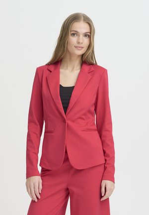 Junge Frau mit langen blonden Haaren, die ein eng anliegendes rotes Sakko und passende Hosen über einem schwarzen Oberteil trägt und vor einem weißen Hintergrund steht.