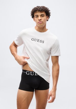 Νέος άνδρας με σγουρά μαλλιά σηκώνει ένα λευκό μπλουζάκι με το λογότυπο GUESS, φοράει μαύρα εσώρουχα τύπου boxer της GUESS, σε απλό ανοιχτόχρωμο φόντο.
