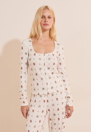 Mujer rubia con camiseta blanca de manga larga y pantalones, ambos con un patrón de pequeñas ilustraciones de perros marrones y verdes, de pie contra una pared neutra.