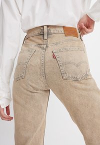 Un jean en denim beige avec une texture décolorée, comportant deux poches arrière et une étiquette en cuir marron à la taille. Les coutures sont visibles.