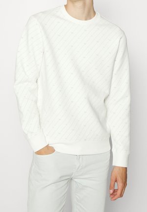 Mann mit weißem Sweatshirt mit dezenten diagonalen Linien und weißer Hose, steht mit einer Hand in der Tasche vor neutralem Hintergrund.