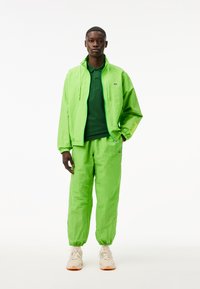 Cazadora verde lima con capucha y puños elásticos, combinada con pantalones a juego. El conjunto cuenta con un polo verde oscuro debajo.