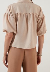 Jasno beżowa bluzka z krótkim rękawem z falbaną z tyłu, wykonana z miękkiego, teksturowanego materiału. Luźny, casualowy krój.