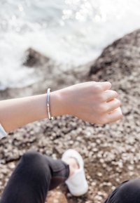 Bracelet en argent sur un poignet, présentant une surface lisse et un design minimaliste, placé sur un sol rocheux avec de l'eau en arrière-plan.