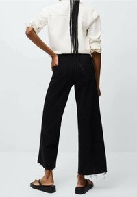 Pantalones negros de pierna ancha con dobladillo deshilachado, combinados con una camisa color crema. Los pantalones cuentan con un diseño clásico de cinco bolsillos y un ajuste relajado.