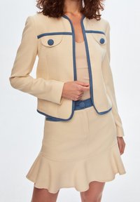 Set a due pezzi in tessuto color crema, con giacca aderente e bordature blu e due tasche frontali, oltre a una gonna a campana coordinata con orlo arricciato.