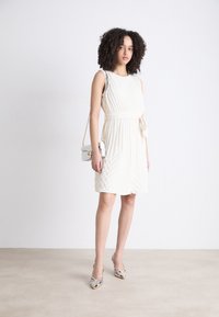 Cream-colored dress with vertical pleats, fitted waist, and bow detail. Paired with silver heeled shoes and a small white handbag.