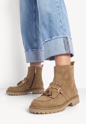 Bottes en daim beige montant à la cheville avec des glands et des boucles, portées avec un jean bleu clair retroussé sur fond blanc.