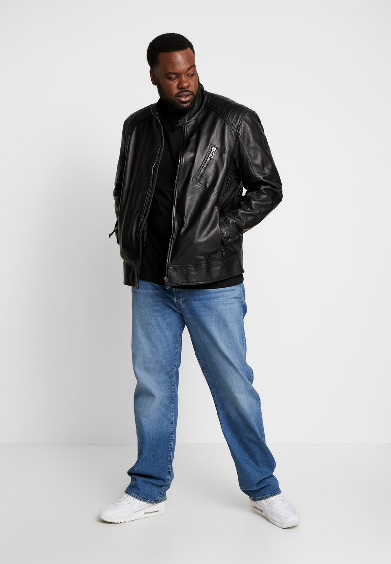 Veste bomber en cuir noir avec des détails matelassés aux épaules et une poche zippée sur la poitrine, associée à un jean bleu clair et des baskets blanches.