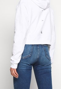 Witte cropped hoodie met een capuchon met trekkoord. Heeft een logo op de achterkant. Gecombineerd met donkerblauwe denim jeans, met een aansluitend ontwerp.