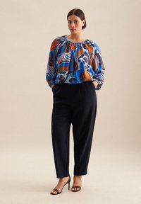 Blusa floreale multicolore con motivi blu, arancioni e marroni; scollo arricciato; pantaloni neri a vestibilità comoda; sandali neri con tacco.