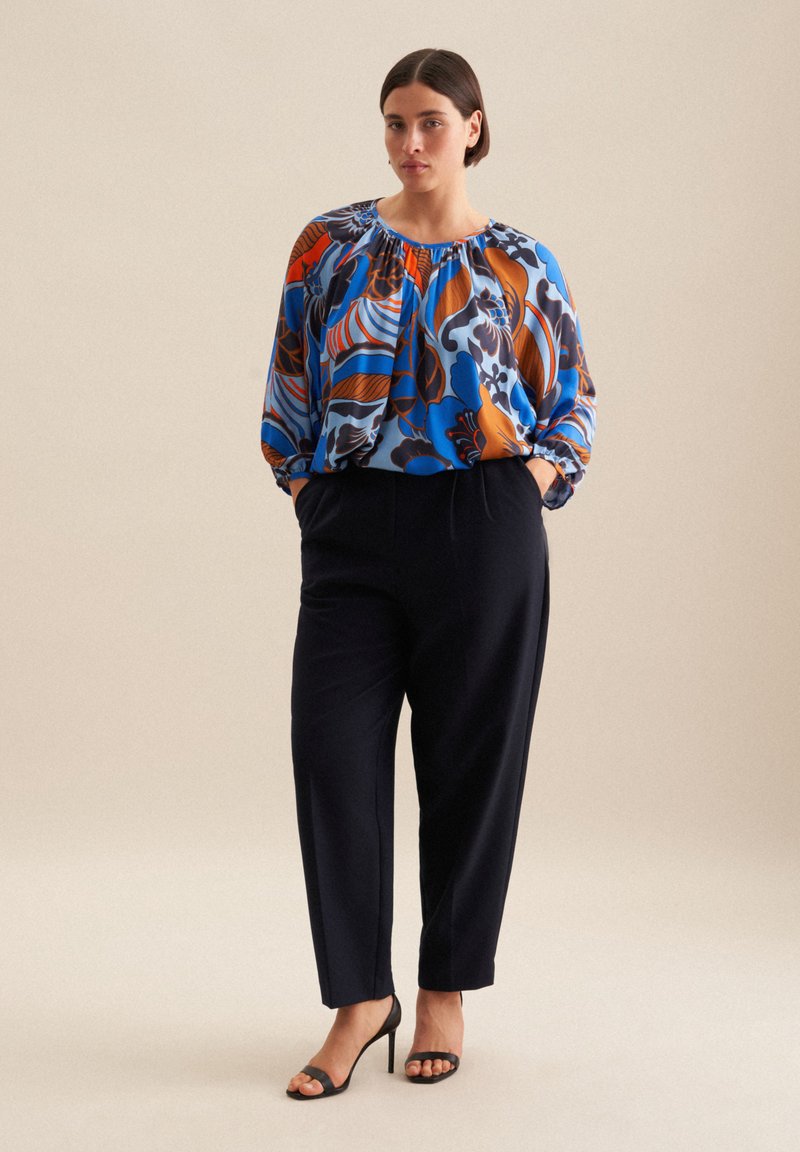 Blusa floreale multicolore con motivi blu, arancioni e marroni; scollo arricciato; pantaloni neri a vestibilità comoda; sandali neri con tacco.