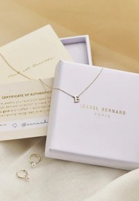 Goldenes Buchstaben-Halsband "B" an einer filigranen Kette, präsentiert in einer weißen Box mit goldener Schrift und einem Echtheitszertifikat. Nebenan befinden sich zwei kleine Perlenohrringe.