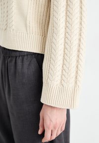 Cream cable-knit sweater with ribbed hem and cuffs, paired with dark gray textured pants. Close-up of sleeve details.